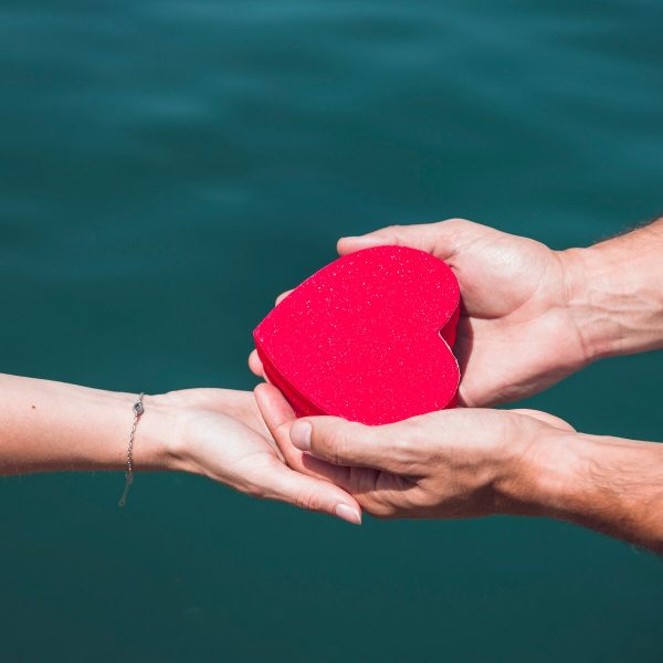 close-up-couple-s-hand-holding-red-heart-shape-front-sea_23-2147887893